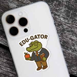 May include: A sticker featuring a cartoon alligator wearing glasses, a suit, and holding an apple and a book. The sticker has the text "EDU-GATOR" in a speech bubble. The sticker is on a white phone case.