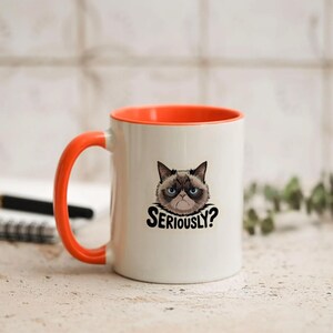 May include: White ceramic mug with an orange interior and handle. The mug features a graphic of a grumpy cat with the text "SERIOUSLY?" in black. The mug is on a light-colored surface.