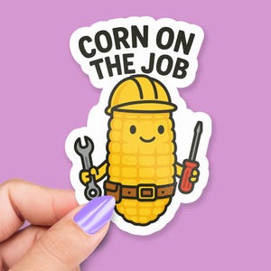 May include: A sticker showing a cartoon ear of corn wearing a yellow hard hat and tool belt, holding a wrench and screwdriver. The text "CORN ON THE JOB" is above the corn character. The sticker is held by a hand with a purple fingernail.