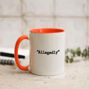 May include: White ceramic mug with an orange interior and handle. The mug features the word "Allegedly" in black text. The mug is on a light-colored surface with a pen and notepad in the background.