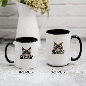 May include: Two white ceramic mugs with black interiors and handles. Each mug features a grumpy cat graphic with the text "Seriously?". The smaller mug is labeled "11oz MUG" and the larger mug is labeled "15oz MUG".