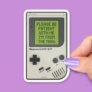 May include: A sticker featuring a retro Nintendo Game Boy. The screen displays the text "PLEASE BE PATIENT WITH ME I'M FROM THE 1900s." The Game Boy is light gray with black buttons and a D-pad. The sticker is held by a hand with a purple painted nail.