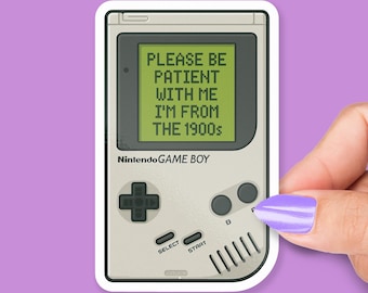 May include: A sticker featuring a retro Nintendo Game Boy. The screen displays the text "PLEASE BE PATIENT WITH ME I'M FROM THE 1900s." The Game Boy is light gray with black buttons and a D-pad. The sticker is held by a hand with a purple painted nail.