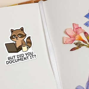 Puede incluir: Una pegatina con un mapache de dibujos animados con un port&aacute;til y una taza. El mapache tiene una expresi&oacute;n de mal humor. El texto de la pegatina dice "BUT DID YOU DOCUMENT IT?" Flores secas en el fondo.