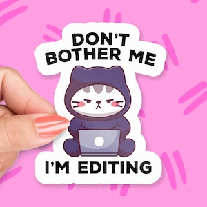 May include: A white sticker with a cartoon cat wearing a purple hooded sweatshirt and using a laptop. The text on the sticker reads "Don't Bother Me I'm Editing."