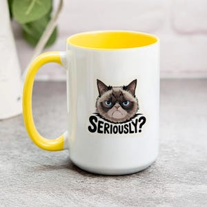 May include: White ceramic mug with a yellow handle and interior. The mug features a graphic of a grumpy cat with the text "SERIOUSLY?" in black. The mug is suitable for hot or cold beverages.