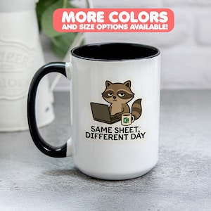 May include: White ceramic mug with a black handle and rim. The mug features a cartoon raccoon with a laptop and a coffee mug, with the text "SAME SHEET, DIFFERENT DAY". A banner at the top reads "MORE COLORS AND SIZE OPTIONS AVAILABLE!"