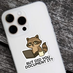 Puede incluir: Una pegatina con un mapache de dibujos animados con un port&aacute;til y una taza de caf&eacute;. El mapache tiene una expresi&oacute;n de mal humor. El texto de la pegatina dice "BUT DID YOU DOCUMENT IT?" La pegatina est&aacute; en una funda blanca para tel&eacute;fono.