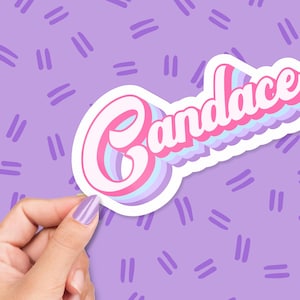 May include: A sticker with the name "Candace" in a retro font, featuring pink, white, and blue layered text with a white outline. The sticker is held against a purple background with a repeating pattern of short, parallel lines.
