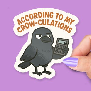May include: A sticker featuring a cartoon crow holding a calculator. The text "ACCORDING TO MY CROW-CULATIONS" is printed in brown above the crow. The crow is gray with a black beak and feet. A hand with purple nail polish is holding the sticker.
