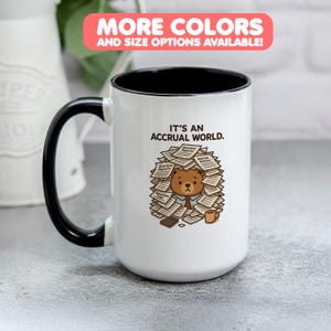 Puede incluir: Taza de cerámica blanca con asa y borde negros. La taza presenta el texto "IT'S AN ACCRUAL WORLD" y un oso de dibujos animados rodeado de papeleo. La parte superior de la taza tiene una pancarta rosa que dice "MORE COLORS AND SIZE OPTIONS AVAILABLE!"