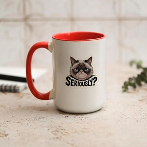 May include: White ceramic mug with a red interior and handle. The mug features a graphic of a grumpy cat with the text "SERIOUSLY?" in black. The mug is designed for hot beverages.