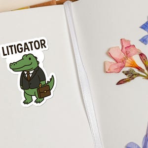 Pode incluir: Um autocolante com um jacar&eacute; de desenho animado vestido com fato e gravata, segurando uma pasta, com a palavra "LITIGATOR" por cima. A imagem tamb&eacute;m inclui flores secas. O autocolante est&aacute; numa superf&iacute;cie branca.