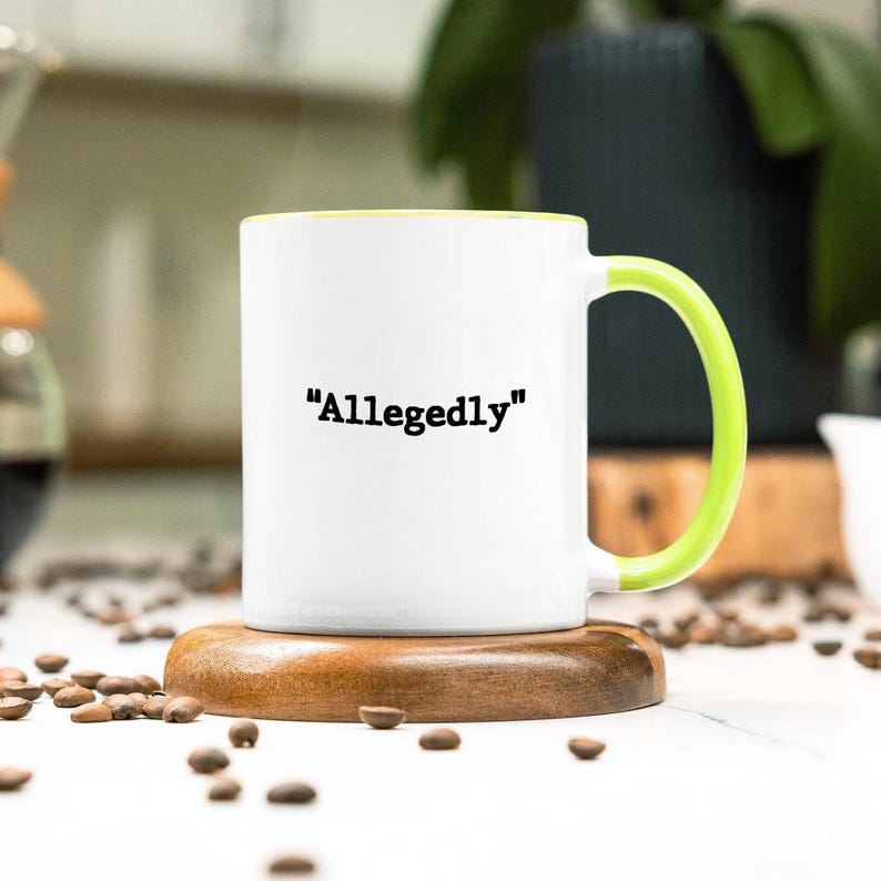 May include: White ceramic mug with the word "Allegedly" in black text. The mug has a bright green handle and interior. The mug is sitting on a round wooden coaster, surrounded by coffee beans.