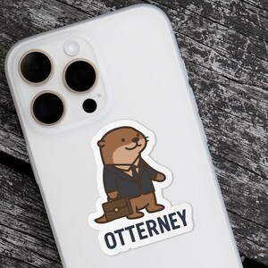 K&ouml;nnte beinhalten: Ein Aufkleber mit einem Cartoon-Otter in einem Anzug, der eine Aktentasche h&auml;lt. Der Otter ist braun und tr&auml;gt einen schwarzen Anzug. Der Text "OTTERNEY" steht darunter in Wei&szlig; mit blauem Rand. Der Aufkleber befindet sich auf einer wei&szlig;en Handyh&uuml;lle.