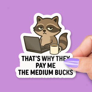 May include: A die-cut sticker featuring a brown and grey cartoon raccoon with a tired expression, sitting at a laptop with a coffee mug. The text below reads, THAT'S WHY THEY PAY ME THE MEDIUM BUCKS, printed in bold, black, sans-serif capital letters.
