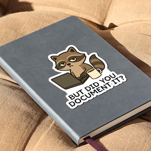 Puede incluir: Una pegatina con un mapache de dibujos animados, un port&aacute;til y una taza de caf&eacute;. El mapache tiene una expresi&oacute;n de mal humor. El texto de la pegatina dice: "BUT DID YOU DOCUMENT IT?" La pegatina est&aacute; sobre un cuaderno gris.