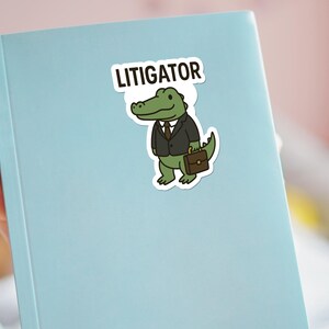 Pode incluir: Um autocolante com um jacar&eacute; de desenho animado de fato e pasta, com a palavra "LITIGATOR" por cima. O autocolante est&aacute; colocado num caderno azul claro.