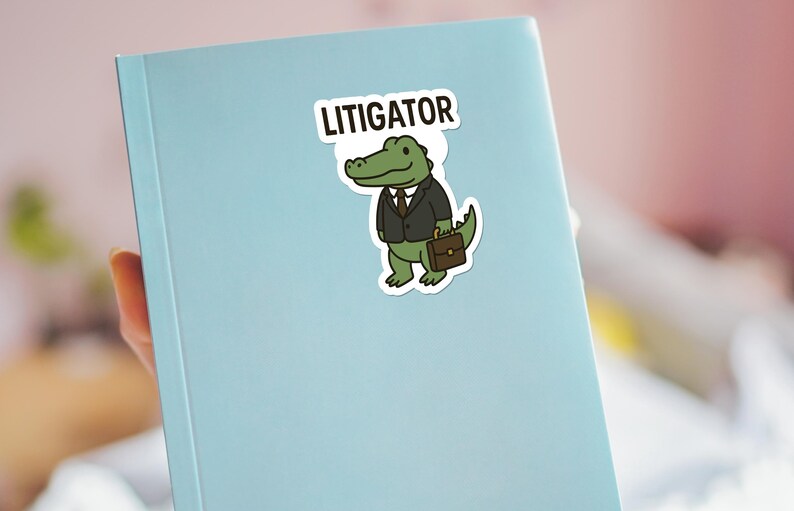 Pode incluir: Um autocolante com um jacar&eacute; de desenho animado de fato e pasta, com a palavra "LITIGATOR" por cima. O autocolante est&aacute; colocado num caderno azul claro.