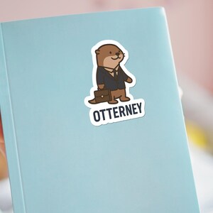K&ouml;nnte beinhalten: Hellblaues Notizbuch mit einem Aufkleber eines Cartoon-Otters, der einen Anzug tr&auml;gt und eine Aktentasche h&auml;lt. Der Aufkleber tr&auml;gt das Wort "OTTERNEY" in wei&szlig;er Schrift auf schwarzem Hintergrund.