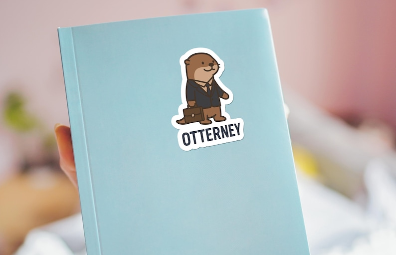 K&ouml;nnte beinhalten: Hellblaues Notizbuch mit einem Aufkleber eines Cartoon-Otters, der einen Anzug tr&auml;gt und eine Aktentasche h&auml;lt. Der Aufkleber tr&auml;gt das Wort "OTTERNEY" in wei&szlig;er Schrift auf schwarzem Hintergrund.