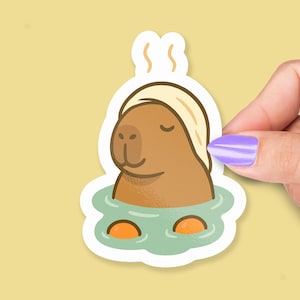 Peut inclure: Un autocollant représentant un capybara de dessin animé se relaxant dans un bain. Le capybara est brun, porte une serviette blanche et est dans un bain vert avec deux tranches d'orange. De la vapeur s'élève du bain.