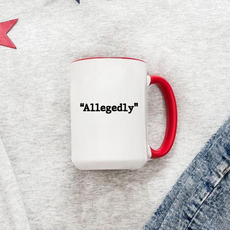 May include: White ceramic mug with a red interior and handle. The mug features the word "Allegedly" in black text. The mug is on a light gray surface with a red star and denim fabric in the background.