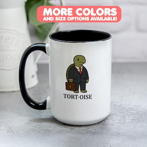 May include: White ceramic mug with a black handle and rim. The mug features a cartoon tortoise in a suit holding a briefcase, with the word "TORT-OISE" below. The top banner reads "MORE COLORS AND SIZE OPTIONS AVAILABLE!"