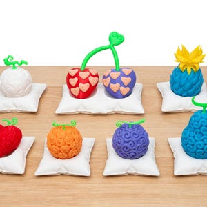 Op de afbeelding: Een verzameling van zeven kleurrijke, getextureerde fruitvormige beeldjes, elk rustend op een klein wit vierkant kussentje. De set bevat diverse vormen en kleuren, zoals een witte bol, rode en paarse harten, een blauwe ananas en gedraaide patronen.