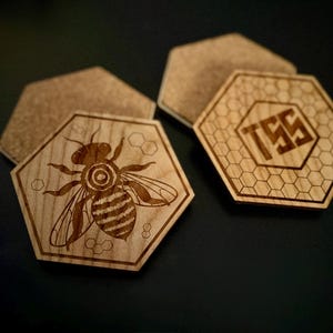 May include: Hexagonal wooden coasters with laser-etched designs. One coaster features a detailed bee illustration, while another displays the letters "TSS" within a honeycomb pattern. The coasters are made of light-coloured wood.