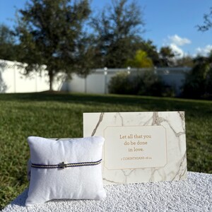May include: A bracelet with a small cross charm on a white pillow, with a card that reads "Let all that you do be done in love." The bracelet has a navy and cream colored band.