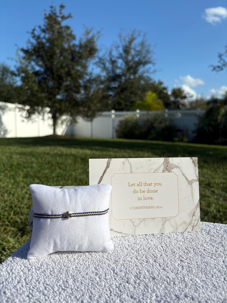 May include: A bracelet with a small cross charm on a white pillow, with a card that reads "Let all that you do be done in love." The bracelet has a navy and cream colored band.