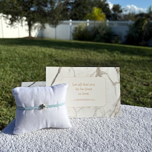 May include: A light blue bracelet with a gold cross charm is displayed on a white pillow. A marble-patterned card with the text "Let all that you do be done in love. 1 CORINTHIANS 16:14" is behind the bracelet. The background is a grassy lawn and a white fence.
