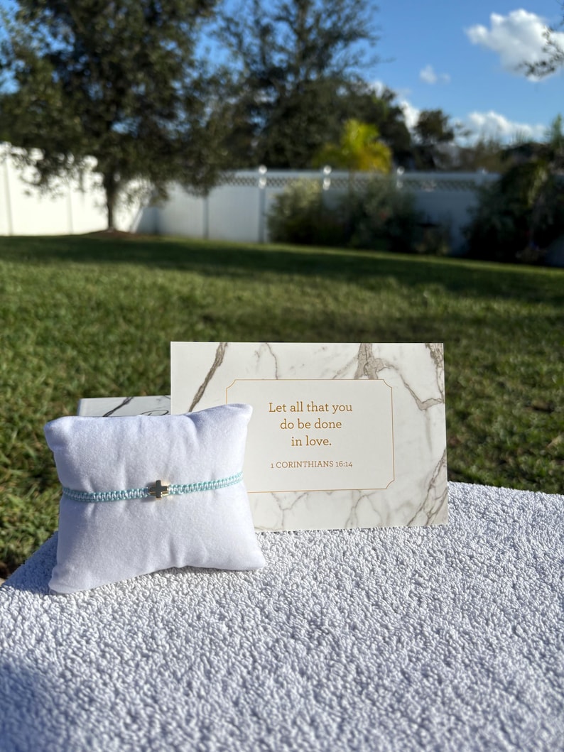 May include: A light blue bracelet with a gold cross charm is displayed on a white pillow. A marble-patterned card with the text "Let all that you do be done in love. 1 CORINTHIANS 16:14" is behind the bracelet. The background is a grassy lawn and a white fence.