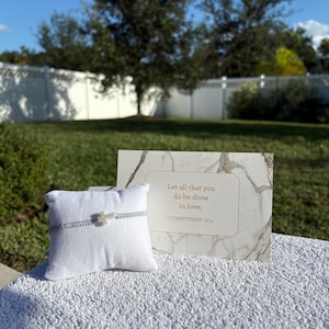 May include: A silver bracelet with a cross charm rests on a white pillow. A marble-patterned card with gold text reads "Let all that you do be done in love." The background features a green lawn and trees.