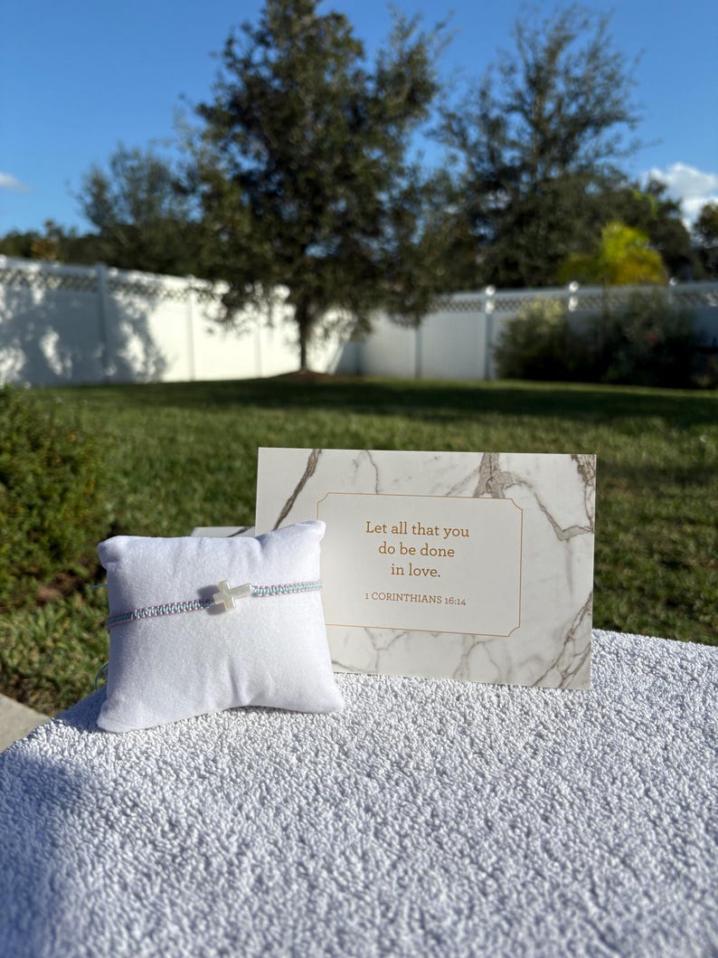 May include: A silver bracelet with a cross charm rests on a white pillow. A marble-patterned card with gold text reads "Let all that you do be done in love." The background features a green lawn and trees.