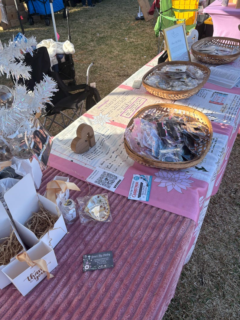 May include: A market stall showcases jewellery and gifts. Wicker baskets contain packaged items, and white boxes with gold ribbon are also present. The table is covered with a pink and white floral patterned tablecloth. A wooden heart is also visible.