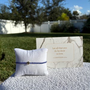 May include: A blue and silver beaded bracelet with a gold cross charm rests on a white pillow. A marble-patterned card with the text "Let all that you do be done in love. 1 CORINTHIANS 16:14" is displayed next to the bracelet.