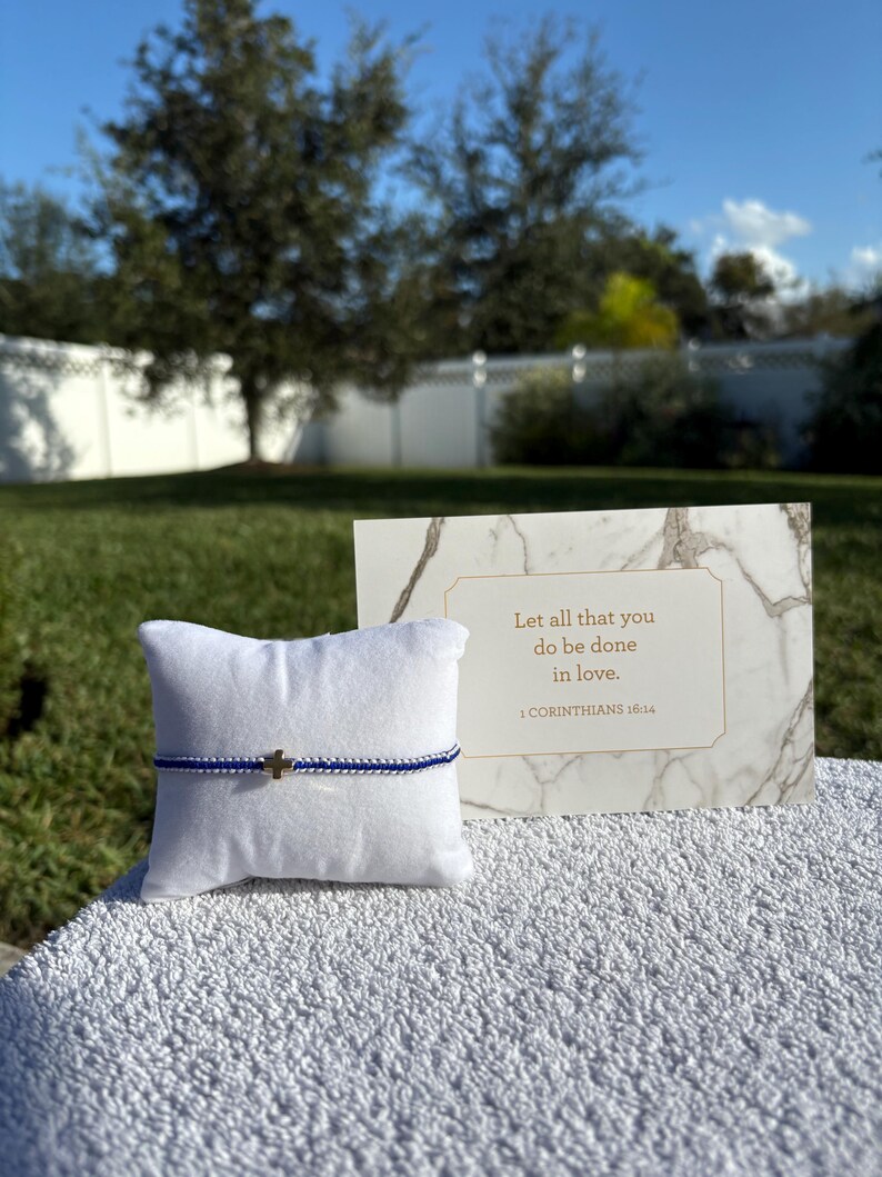 May include: A blue and silver beaded bracelet with a gold cross charm rests on a white pillow. A marble-patterned card with the text "Let all that you do be done in love. 1 CORINTHIANS 16:14" is displayed next to the bracelet.