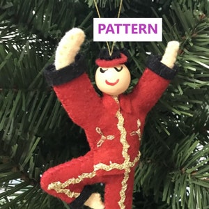 May include: A felt Christmas ornament depicting a person in a red and black outfit with gold trim. The ornament is hanging on a green Christmas tree. The ornament has a smiling face and is in a dancing pose. The ornament has the word "PATTERN" written on it.