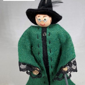 May include: A felt doll wearing a green robe with black trim and a black witch's hat with a feather. The doll has a painted face with glasses and is holding a black wand.