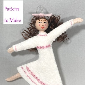 May include: A felt doll ornament with brown curly hair, a pink bow, and a white dress with pink trim. The doll is in a dancing pose with one leg raised.