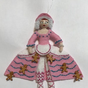 May include: A pink felt Christmas ornament of a gingerbread woman wearing a white apron and a pink dress with blue trim. She has a white felt hat and is surrounded by gingerbread men.