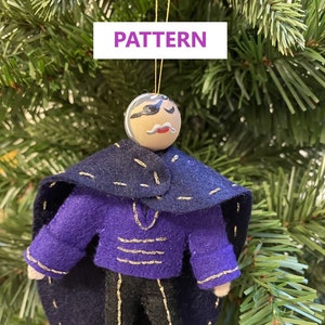 以下が含まれることがあります： 紫色のジャケットに金の縁取り、黒いマント、黒いズボンを着用した人物のフェルト製オーナメント。オーナメントは、口ひげと黒い眼帯が描かれた顔をしています。オーナメントはクリスマスツリーの紐にぶら下がっています。