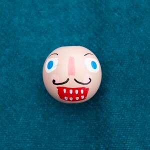 May include: A whimsical, hand-painted wooden ornament shaped like a face. The ornament features blue eyes, a pink nose, a black mustache, and a red mouth with white teeth. The background is a teal fabric.