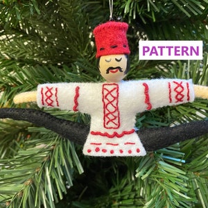 May include: A felt ornament of a person wearing a red hat and white embroidered shirt with black pants. The ornament is in a split position with arms outstretched and legs spread apart. The ornament is hanging on a green Christmas tree.