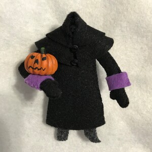 Puede incluir: Una figura de fieltro de un personaje sin cabeza que viste un abrigo negro con tres botones negros y puños morados. El personaje sostiene una pequeña calabaza naranja con una cara de jack-o'-lantern pintada a mano en color negro.