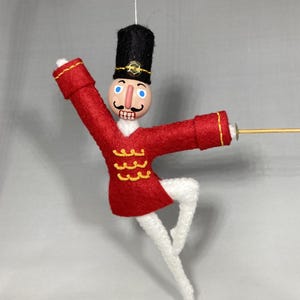 May include: A handcrafted Christmas ornament depicting a toy soldier in a ballet pose. The soldier wears a red felt jacket with gold trim, white tights, and a black hat. The ornament is suspended by a thin string.