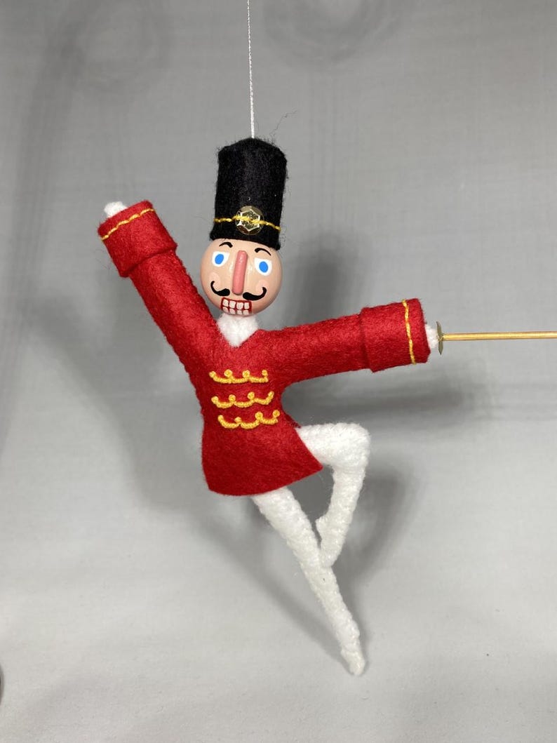 May include: A handcrafted Christmas ornament depicting a toy soldier in a ballet pose. The soldier wears a red felt jacket with gold trim, white tights, and a black hat. The ornament is suspended by a thin string.
