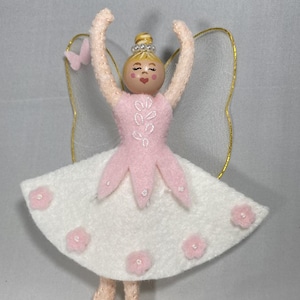 Op de afbeelding: Handgemaakt feeënornaat met een roze lijfje en een witte rok versierd met roze bloemen. De fee heeft een houten hoofd, opgeheven armen en gouden vleugels. Een kleine roze vlinder is aan de vleugel bevestigd. Ideaal voor een kinderkamer of als feestelijke decoratie.